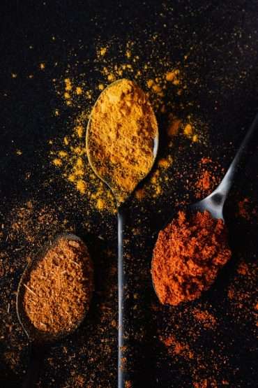 Tablespoons with dry condiments spilled on black background